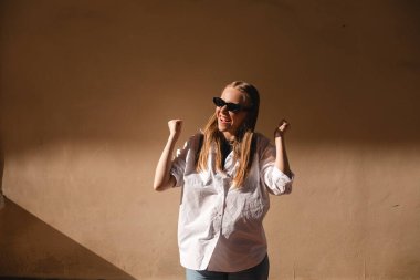 Neşeli, güzel bir kadın kutlama yapıyor, inançsızlık ve heyecanla ödül kazanıyor, şampiyon gibi tezahürat yapıyor, iyi haberlerden bağırıyor ve zafer kazanıyor, dışarıda bej duvarın yanında duruyor..