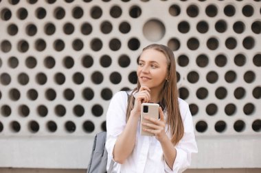 Rüya gören genç bir kadın düşünün duvarın yanında poz veriyor şehir arka planında cep telefonu kullanarak..