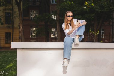Genç şık sarışın kadın akıllı telefonuyla mesaj yazıyor Green City Park 'ta oturuyor, hippi turistler şehirde geziyor ve cep telefonuyla seyahat planlıyor. İleti okunurken gülümse.