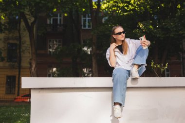Güzel sarışın kadın kamerada selfie çekiyor. Şirin kız parkta oturur ve kameraya bakar. Doğada, ince kız kamerayla flört eder. Kız beyaz gömlek, kot pantolon ve spor ayakkabı giyer..