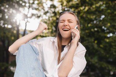 Gülümseyen genç kız internette cep telefonuyla geziyor. Günlük kıyafetleri içinde, kazanan gibi el kol sallıyor. Sarışın kadın mutlu görünüyor, zaferi, zaferi, başarıyı kutluyor. Kızın cep telefonu var..