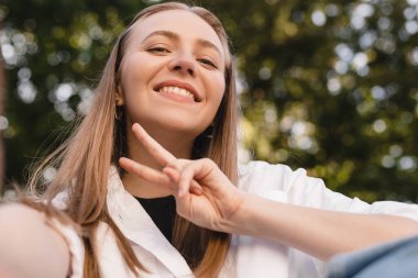Düşünceli sarışın kadın arka planda poz vererek selfie çekiyor. Mutlu iş kadınının açık hava çekimi barış işareti. İş özgürlüğü tarzı. Kız elini kameraya kaldırır, dişlek gülümsemesini gösterir,.