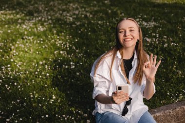 Gülümseyen genç kadın cep telefonunu tut ve tamam işareti göster. Yeşil tarlalarda parktaki çiçeklerle otururken sağ el hareketi yap. Anlaştık, onaylandı, onaylandı. Sarışın kadın beyaz gömlek giyiyor..