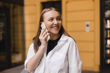 Sarı duvar binasının önünde akıllı telefondan konuşan mutlu kadın. Güzel, şık bir kadın telefonda konuşuyor, cep telefonuyla dışarıda yürüyor. Kız yana bak..