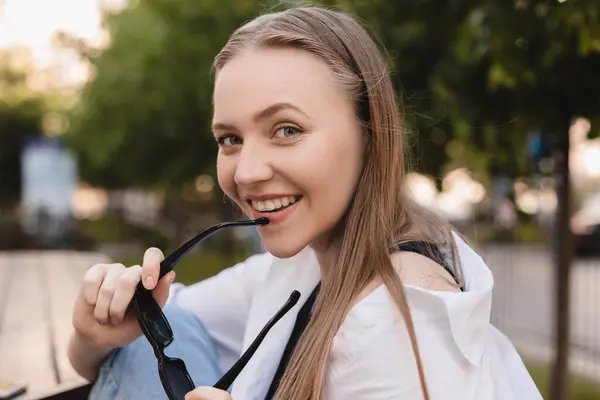 Beyaz gömlekli dalgın sarışın kadın siyah gözlük taktı ve sokak arkasında poz verdi. Şehir parkında oturan mutlu hippi kadının açık havada çekilmiş fotoğrafı. Boho özgürlük tarzı.