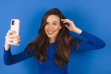 Esmer kız telefona bakıyor ve saçını düzeltiyor. Kadın telefonu ayna gibi kullanıyor. Genç kız mavi arka planda telefonla fotoğraf çekiyor..