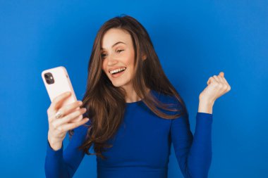 Fun young woman hold in hand use mobile cell phone doing winner gesture clenching fists isolated on blue background studio. Girl using smartphone apps winning online celebrating discount gift voucher clipart