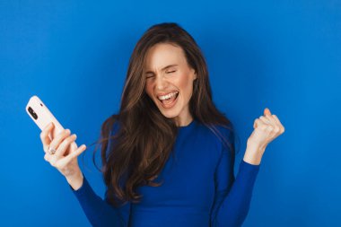 Eğlenceli genç kadın cep telefonu kullanıyor. Mavi arka plan stüdyosunda izole edilmiş yumruklarını sallıyor. Akıllı telefon uygulaması kullanan kız internette indirim kuponu kazanıyor.