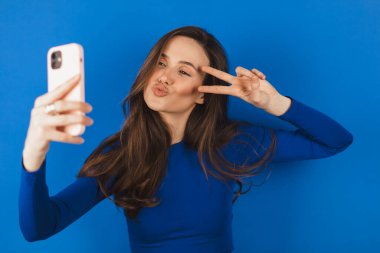 Güzel genç esmer kadın cep telefonunu elinde tutuyor, selfie çekiyor, sakso çekiyor, barış işareti ile poz veriyor, mutlu gülümsüyor, fotoğraf çekiyor, mavi arka planda poz veriyor..
