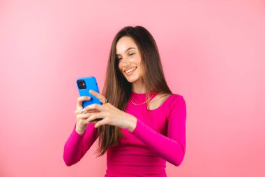 Gülümseyen esmer kadın cep telefonu kullanıyor. Mutlu mesajlaşma, sohbet, sosyal ağlarda mesajlaşma. Pembe arka planda izole edilmiş modern teknolojilere bağımlı. Çevrimiçi iletişim.