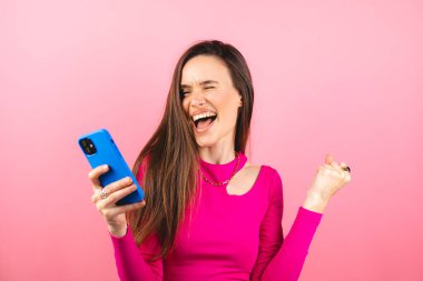 Eğlenceli genç kadın cep telefonu kullanıyor. Pembe arka plan stüdyosunda izole edilmiş yumruklarını sallıyor. Akıllı telefon uygulaması kullanan kız internette indirim kuponu kazanıyor..