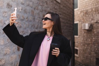 Mutlu genç bir kadın cep telefonuyla kendi fotoğrafını çekiyor. Kadın şehirde akıllı telefonuyla selfie çekiyor. Esmer saçlı kadın dışarıda fotoğraf çekiyor. Siyah gözlük takıyor, kahve içmeye gidiyor..