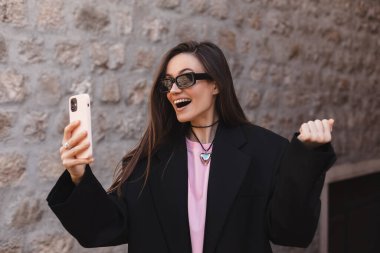 Mutlu gülümseyen genç esmer kadın heyecanlı yüz ifadesiyle akıllı telefonu tutuyor, oynadığına, piyangoda kazandığına, iyi haberler okuduğuna inanamıyor, falına inanamıyor. Gözlük takan kız.
