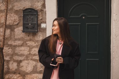 Modaya uygun kıyafetleri olan mutlu güzel bir kadın sokakta yürüyor ve keyfi yerinde olsun diye elinde fincanda kahve tutuyor. Siyah ceketli kız mutlu görün ve yeşil kapının yanına bak..