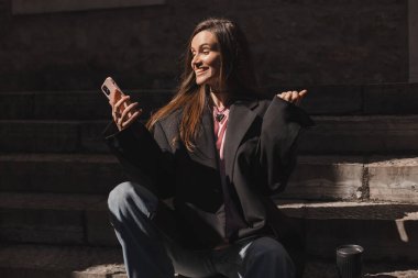 Sokakta siyah takım elbiseli, mutlu cep telefonu tutan, gülümseyen, jest kazanan bir kadın. Esmer kadın mesajları okuyor ve mutlu görünüyor. Zaferi kutluyor, başarıyı..