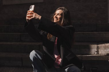 Güzel, güler yüzlü, modaya uygun bir iş kadını eski kasabanın merdivenlerinde oturuyor ve selfie çekiyor, cep telefonunu tutuyor, mutlu görünüyor. Harika bir kadın güneşli bir günde mutlu görünür..