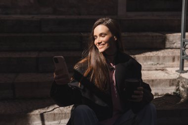 Genç şık esmer kadın akıllı telefondan mesaj yazıyor. Eski şehir caddesinde yürüyor, hipster turistler seyahat ediyor ve cep telefonu kullanıyor. Mesaj okunduğunda mutlu görün, merdivenlerde otururken..