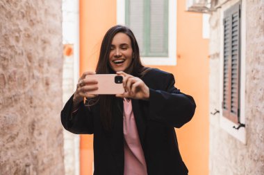 Güzel genç bir kadın cep telefonu kullanarak fotoğraf çekiyor. Esmer kadının portresi siyah ceket giyer dışarı çıkar ve akıllı telefondan fotoğraf çeker..