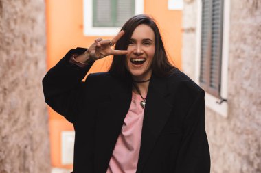 Siyah ceketli havalı esmer kadın, pembe uzun kollu üst ve kalp şeklinde kolyesi V işareti, barış ve dostluk işareti, ağzı açık ve dışarıda poz veriyor. Mutlu kadın şehirde yürüyüp poz veriyor..