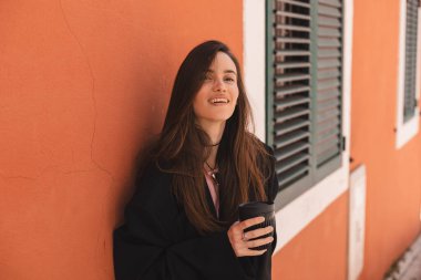 Turuncu duvarın yanında dikilen, elinde fincanla gezen mutlu bir kadın kahve ya da çayla gider. Kadınlar sokakta dinlenirken cappuccino içer..