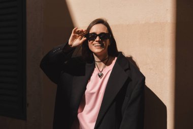 Esmer, çekici bir kadının portresi güneş gözlüğü takıyor ve bej duvarın yanında güneş ışığıyla gülümsüyor. Siyah ceket, gözlük, pembe uzun kollu üst ve kalp şeklinde kolye takan bir kız..