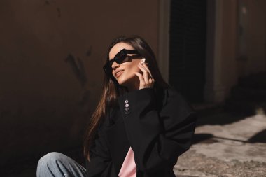 Merdivenlerde oturan ve kablosuz kulaklıklara dokunan gülümseyen genç bir kadın. Kadın eski şehirde müzik dinleyerek rahatlıyor. Teknoloji kullanan genç bir kadın, siyah gözlük takıyor, siyah ceket giyiyor, cep telefonu kullanmıyor..