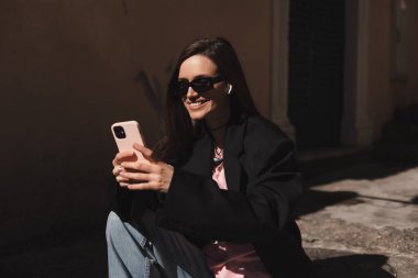 Genç şık esmer kadın akıllı telefondan mesaj yazıyor. Eski şehir caddesinde yürüyor, hipster turistler seyahat ediyor ve cep telefonu kullanıyor. Mesaj okunduğunda mutlu görün, merdivenlerde otururken..