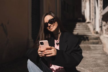 Genç şık esmer kadın akıllı telefondan mesaj yazıyor. Eski şehir caddesinde yürüyor, hipster turistler seyahat ediyor ve cep telefonu kullanıyor. Mesaj okunduğunda mutlu görün, merdivenlerde otururken..