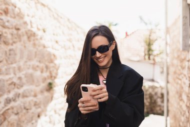 Şehir sokağının dışında gülümseyen modern aletleri tutan çekici genç bir bayanın fotoğrafı. Düşünceli esmer kadın serbest çalışan, siyah ceket giyen, eski şehirde yürüyen, telefonu elinde tutan..