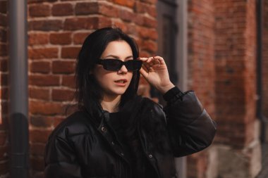 Siyah kabarık ceketli dalgın esmer kadın siyah gözlük takmış ve ofis arka planında poz vermiş. Gülümseyen mutlu iş kadınının açık hava fotoğrafı. Şehvetli görün..