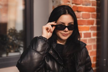 Siyah kabarık ceketli dalgın esmer kadın siyah gözlük takmış ve ofis arka planında poz vermiş. Gülümseyen mutlu bir iş kadınının açık havada çekilmiş fotoğrafı..