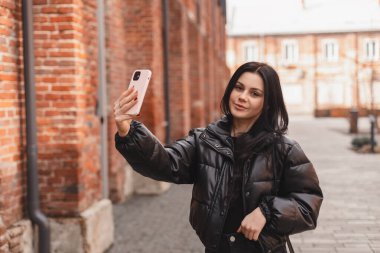 Düşünceli esmer kadın sokak tuğlası bina arka planında poz vererek selfie çekiyor. Açık havada çekilen mutlu hippi bayan görüntüsü video çağrısı yapıyor, gülümsüyor. Siyah kabarık ceket giyen kız kamerasını kaldırıyor..