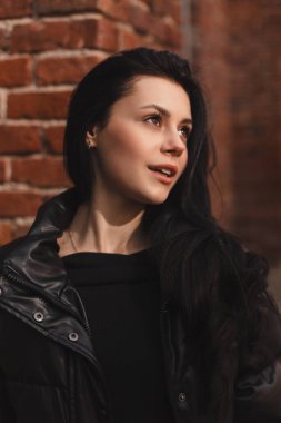 Tuğla duvarın yanında gülümseyen güzel kadın. Gülümseyen esmer bir kızın açık hava portresi. Mutlu neşeli kız şehvetli görünüyor, şehirde hayaller kuruyor, moda gibi görünüyor. Siyah bir kabarık ceket giyiyor..