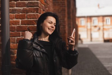 Mutlu gülümseyen genç bir kadın heyecanlı bir yüz ifadesiyle akıllı telefonu tutuyor, oynadığına, piyangoda kazandığına, iyi haberler okuduğuna inanamıyor, falına inanamıyor. Siyah kabarık ceket giyen bir kız.