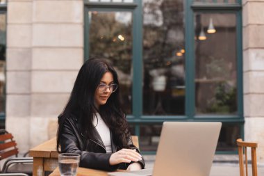 Öğrenci kadın, kafede dizüstü bilgisayar kullanarak deri ceket ve gözlük takıyor. Bir restoranda oturmuş bilgisayarla çalışan gülümseyen güzel bir kız. Açık kafede internette toplanan genç bir kadın..