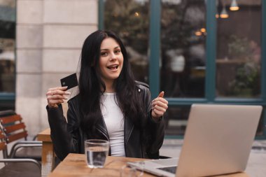 Kredi kartı tutan ve dizüstü bilgisayar kullanan genç bir kız. Kafede para ödeyen bir kadın. Çevrimiçi alışveriş, e-ticaret, internet bankacılığı, harçlık, restoran konseptiyle çalışmak. Kız baş parmağını kaldır..
