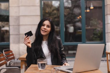 Kredi kartı tutan ve dizüstü bilgisayar kullanan genç bir kız. Kafede para ödeyen bir kadın. Çevrimiçi alışveriş, e-ticaret, internet bankacılığı, harcama, restoran konseptinden çalışma.