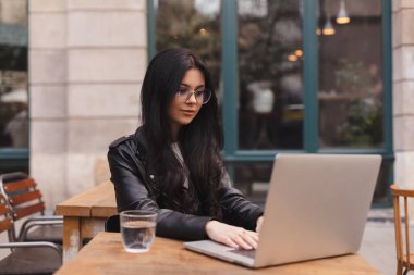 Öğrenci kadın, kafede dizüstü bilgisayar kullanarak deri ceket ve gözlük takıyor. Bir restoranda oturmuş bilgisayarla çalışan gülümseyen güzel bir kız. Açık kafede internette toplanan genç bir kadın..
