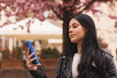 Çekici esmer bayan öğrenci siyah deri ceket giyiyor cep telefonunu tutuyor ve sohbet ediyor, mesajlaşıyor, mutlu görünüyor ve gülümsüyor. Çiçeklerin yanında yürüyen inanılmaz eğlenceli kadın..