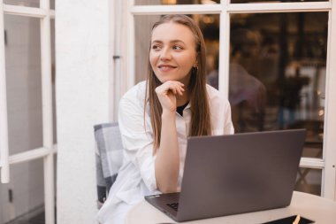 Genç, sarışın, neşeli, neşeli bir kadın laptopla birlikte açık kafede çalışırken gülümsüyor, çalışıyor ve dinleniyor, başını tutuyor, düşünceli görünüyor ve hayal kuruyor..