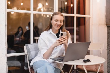 Çekici sarışın kadın dışarıda kafede oturuyor, dizüstü bilgisayarla çalışıyor, cep telefonuna bakıyor, sohbet ediyor, mesajlaşıyor, akıllı telefon kullanan mutlu kadın, kablosuz kulaklık takıyor ve beyaz gömlek giyiyor..