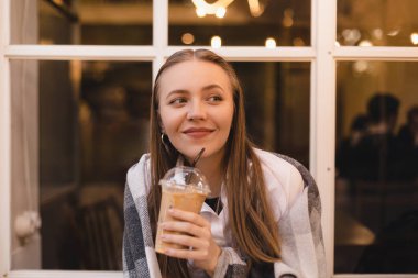 Kafede oturan mutlu, gülümseyen sarışın kadın, buzlu lattenin tadını çıkarıyor, ekoseli gömleğini örtüyor. Hayalperest ve düşünceli görün, akşamları işten sonra kahve molası verin..