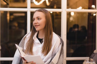 Mutlu, rüya gibi, genç bir kadının kitap okuduğu, hayal kurduğu ve kafeteryada veya restoranda otururken düşünceli göründüğü bir fotoğraf. Rüyamda sarışın kadın kitapla dinleniyor. Kadının omuzları ekoseyle kaplı..