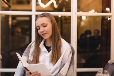 Mutlu, rüya gibi, genç bir kadının kitap okuduğu, hayal kurduğu ve kafeteryada veya restoranda otururken düşünceli göründüğü bir fotoğraf. Rüyamda sarışın kadın kitapla dinleniyor. Kadının omuzları ekoseyle kaplı. Kitaba odaklan