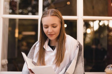 Mutlu, rüya gibi, genç bir kadının kitap okuduğu, hayal kurduğu ve kafeteryada veya restoranda otururken düşünceli göründüğü bir fotoğraf. Rüyamda sarışın kadın kitapla dinleniyor. Kadının omuzları ekoseyle kaplı. Kitaba odaklan