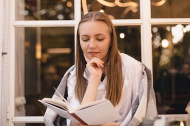 Mutlu, rüya gibi, genç bir kadının kitap okuduğu, hayal kurduğu ve kafeteryada veya restoranda otururken düşünceli göründüğü bir fotoğraf. Rüyamda sarışın kadın kitapla dinleniyor. Kadının omuzları ekoseyle kaplı..