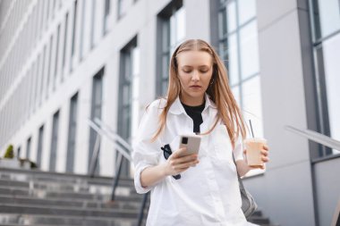 Kendine güvenen genç bir iş kadını elinde cep telefonu, bardak, kahve fincanı, modern ofis binasının önünde cep telefonuyla mesajlaşıyor. Çok meşgul bir kadın..