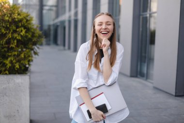 Sarı saçlı iş kadını ofis binasının önünde poz verirken dizüstü bilgisayarını, cep telefonunu ve kitabını kucaklıyor. Hayalindeki iş adamı dışarıda gülüyor ve çenesini tutuyor..