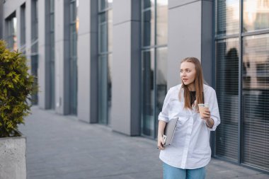 Güzel bir kadın, ofis binasının yakınında buzlu latte ile işe gidiyor. Başarılı bir iş kadını elinde bir fincan sıcak içecek, dizüstü bilgisayar, işe giderken yolda bir telefon tutuyor. Yüksek çözünürlük.