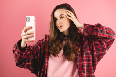 Esmer kız telefona bakıyor ve saçını düzeltiyor. Kadın telefonu ayna gibi kullanıyor. Pembe arka planda telefonla fotoğraf çeken genç bir kız..